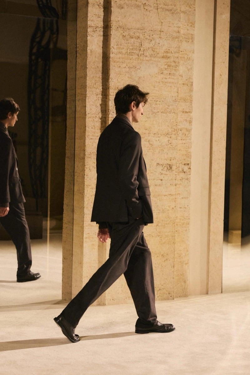Massimo Dutti Limited Edition fall-winter 2026: Side view of a model walking in a dark tailored jacket and wide trousers.