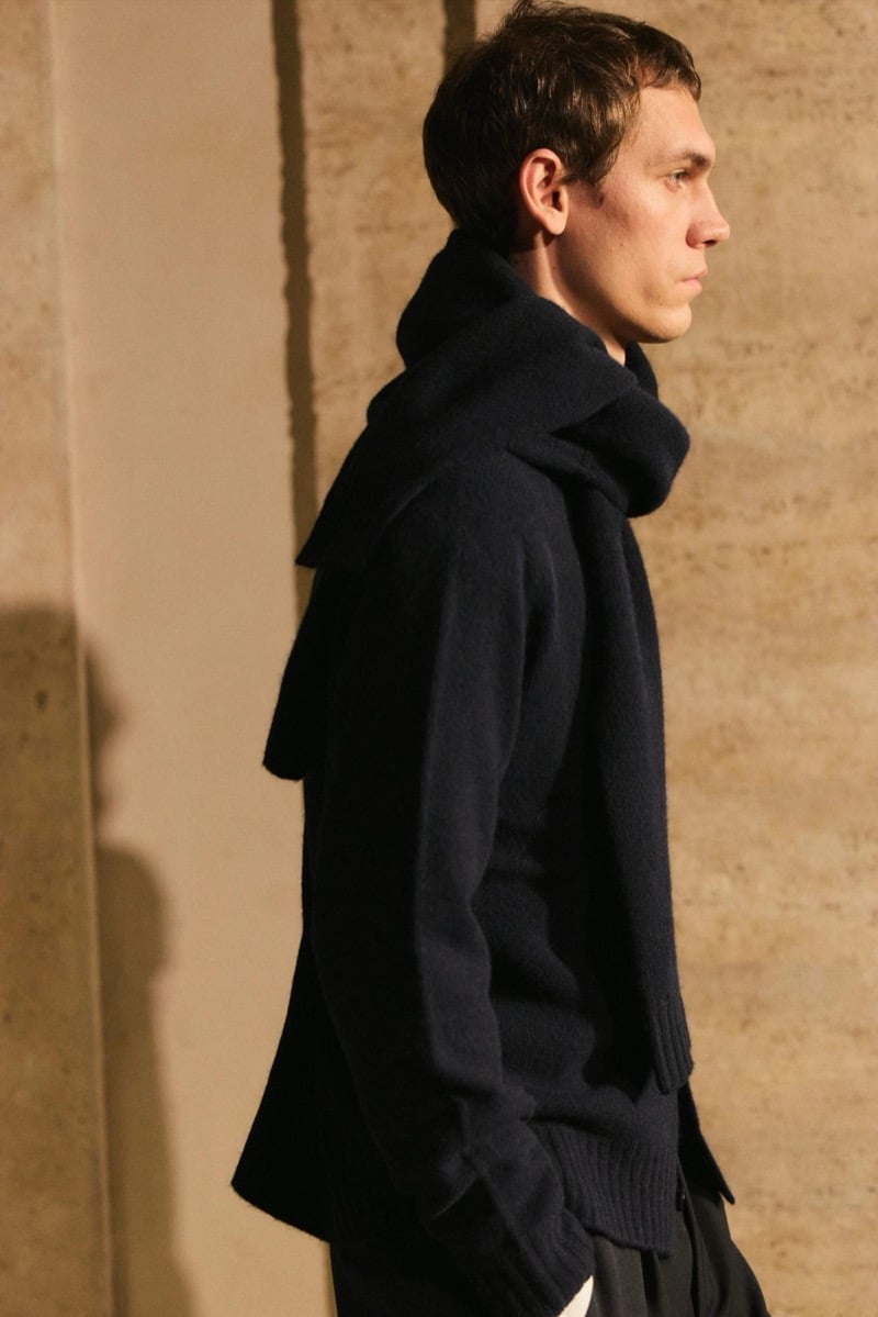 Massimo Dutti Limited Edition fall-winter 2026: Side view of a model wearing a dark sweater and scarf with pleated trousers.