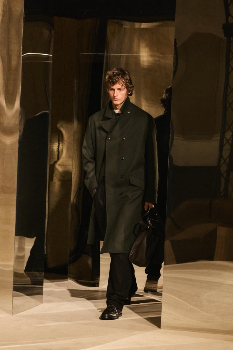 Massimo Dutti Limited Edition fall-winter 2026: Model walks in an olive double-breasted overcoat with dark trousers, carrying a bag.