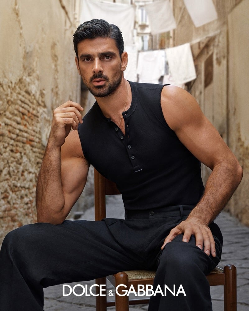 Michele Morrone seated on a wooden chair in a ribbed sleeveless henley and wide trousers, one hand raised near his face, in a Naples alley