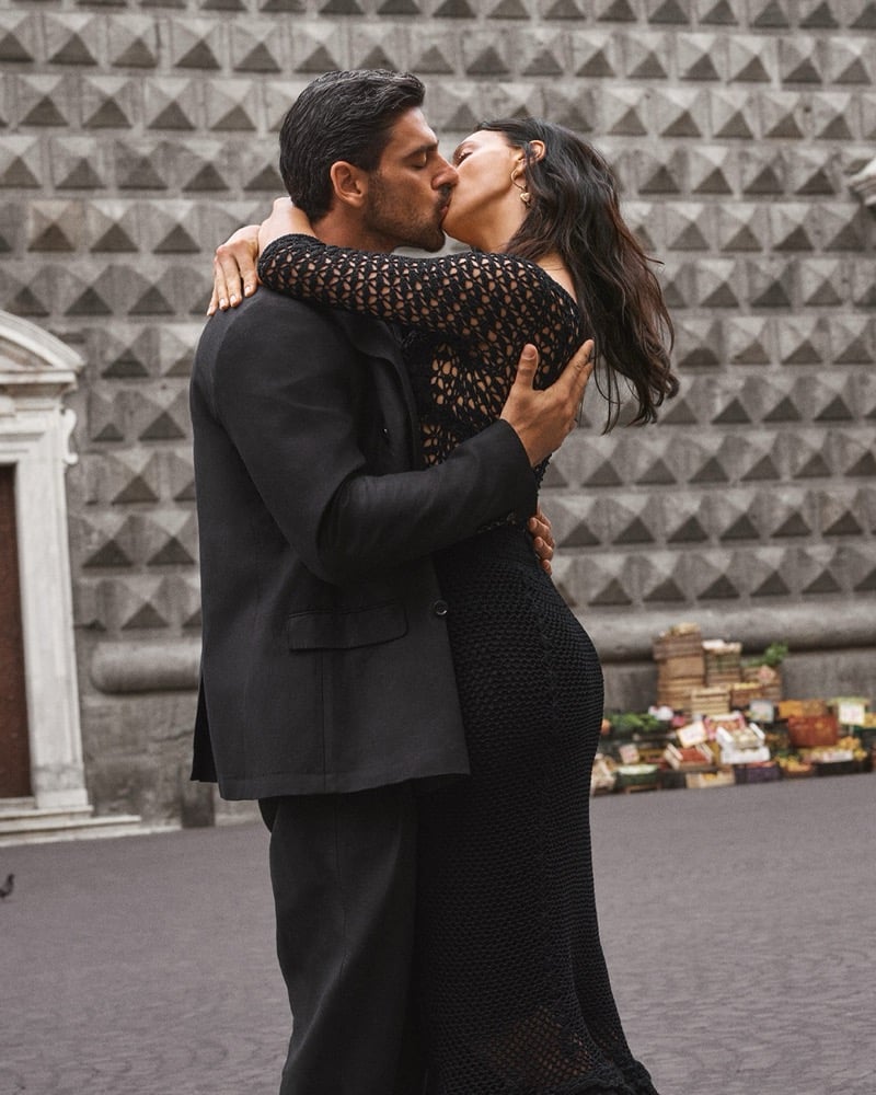 Michele Morrone in a dark jacket kissing Irina Shayk in a crochet dress before a diamond-cut stone facade and a produce stand in a Naples piazza