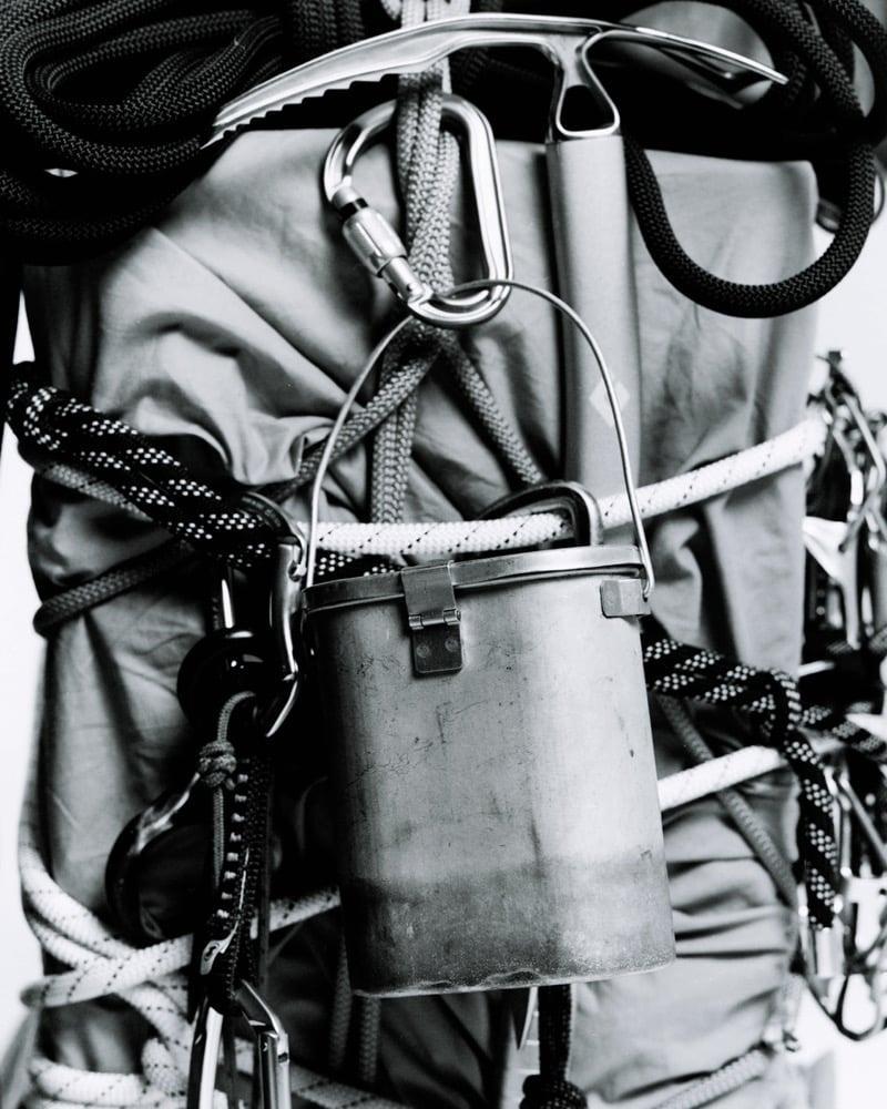 Black-and-white still life of an ice axe, a carabiner, and a metal pot hanging over coiled ropes lashed to a backpack