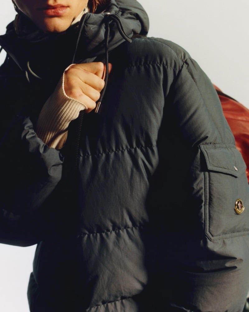 Close-up of a model in a dark hooded puffer jacket with a Moncler patch on the chest pocket, one hand gripping the drawcord at the collar