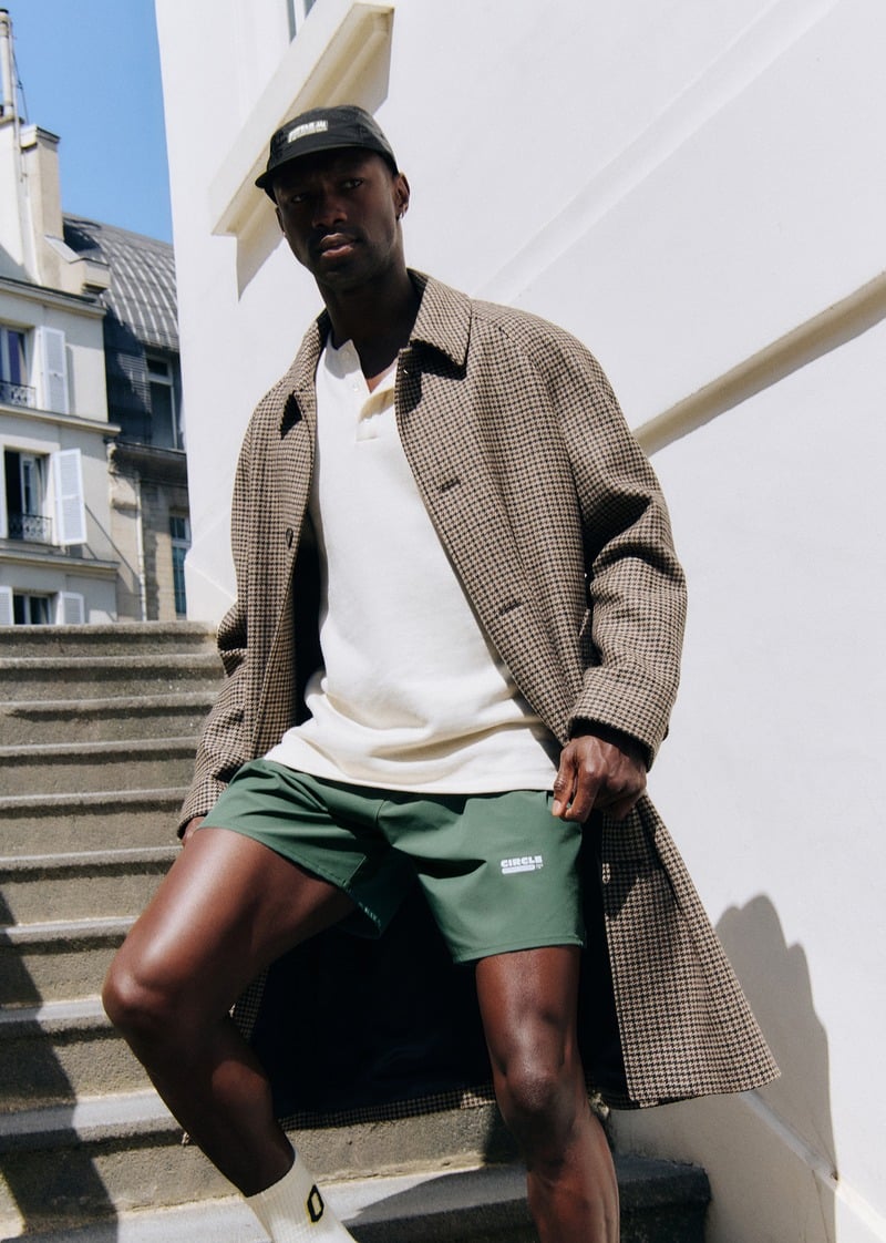 Sadiq Desh wearing a checked overcoat, pale polo shirt, green running shorts and black cap on outdoor steps