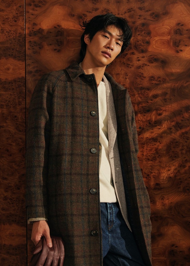 Sanggun Lee leans against a wood-paneled wall in a checked Mikkelsen coat and white shirt.