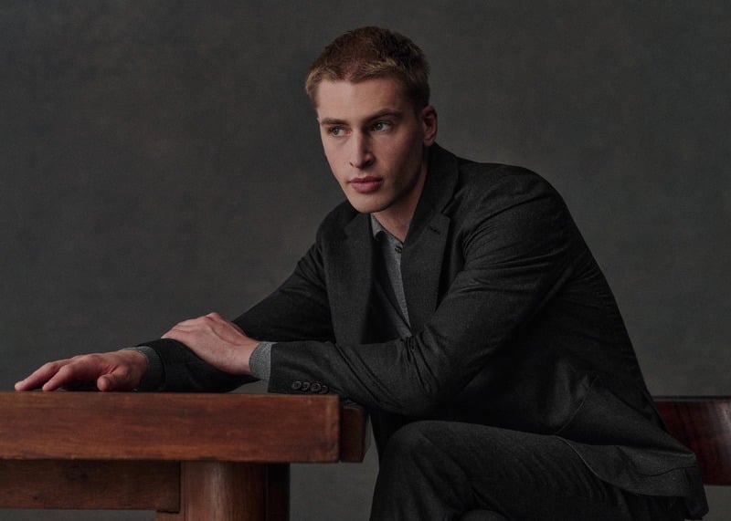 Max Turland leans across a wooden table in a charcoal Riddle jacket and patterned shirt.
