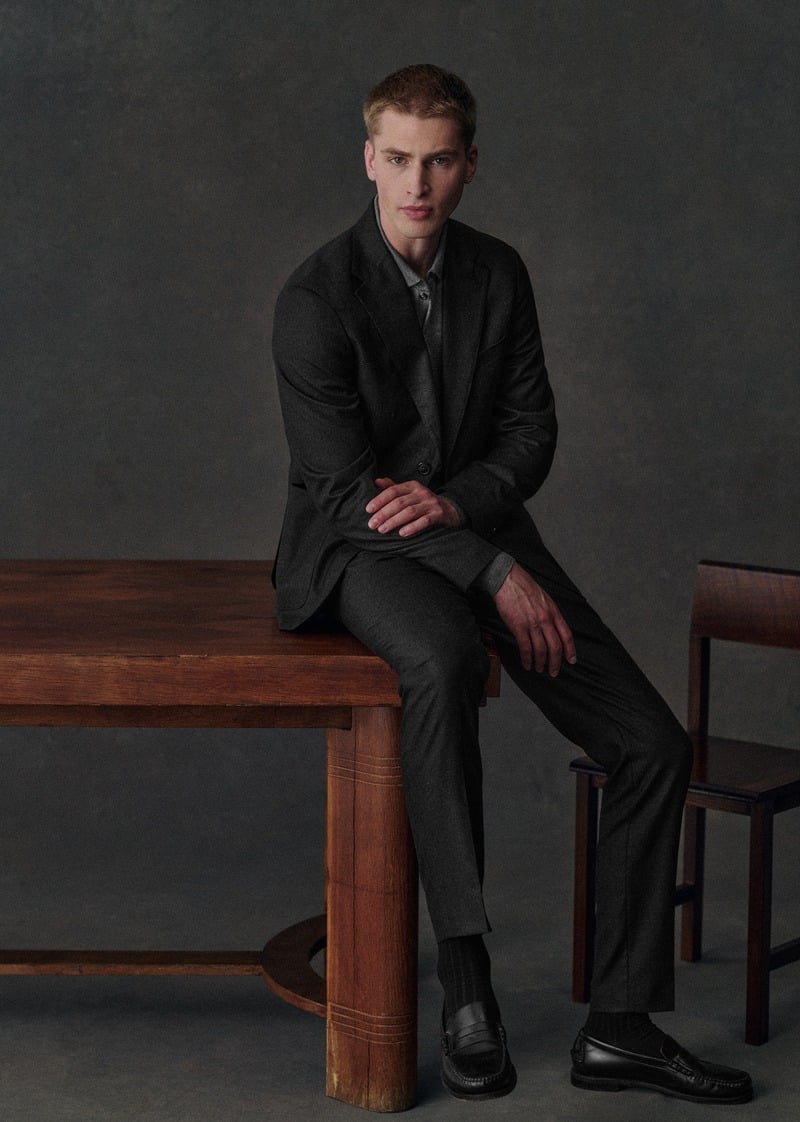 Max Turland sits on a wooden table in the charcoal Riddle jacket and Carl trousers with black shoes.