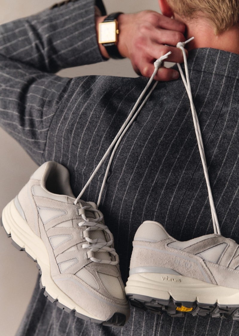 Max Turland holds white-beige OE10 sneakers against a pinstripe outfit.