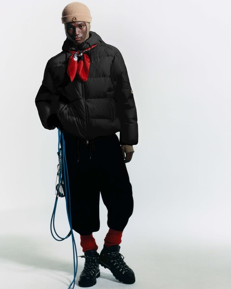Olu Charles in a tan knit beanie, a black hooded puffer jacket, a red neckerchief, red socks, and black lace-up hiking boots, a blue climbing rope hanging at his side