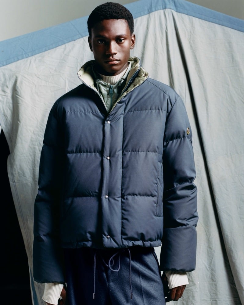 Olu Charles in a boxy blue quilted jacket over a pale green zip layer and a ribbed knit collar, standing in front of a blue-trimmed canvas tent