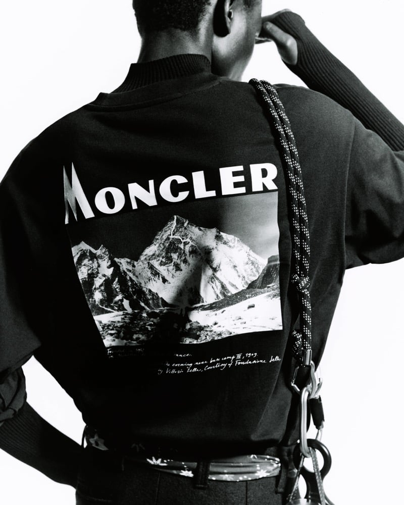 Black-and-white back view of Olu Charles in a Moncler sweatshirt with a mountain photograph and logo printed across the shoulders, a rope and carabiners over one shoulder