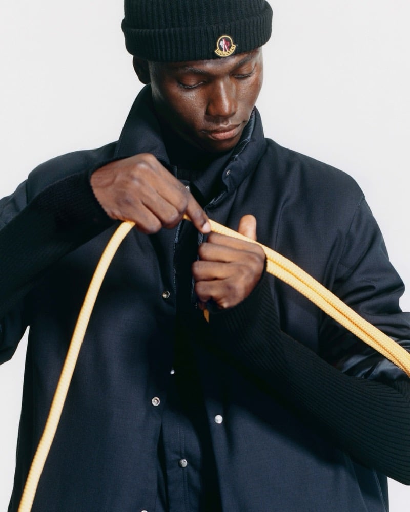 Olu Charles looking down in a knit beanie with a Moncler patch and a dark snap-front jacket with ribbed sleeves, pulling a yellow climbing rope through his hands