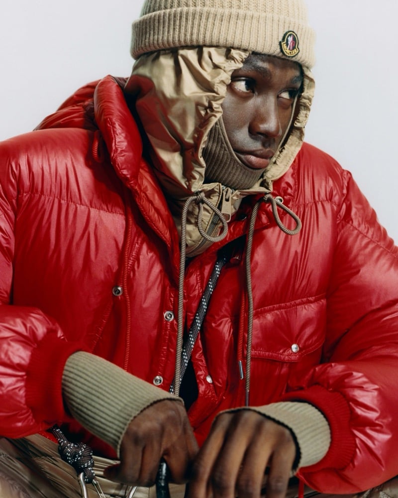 Olu Charles glancing aside in a glossy red Moncler puffer jacket, a tan knit beanie, and a tan quilted hood with rib-knit cuffs