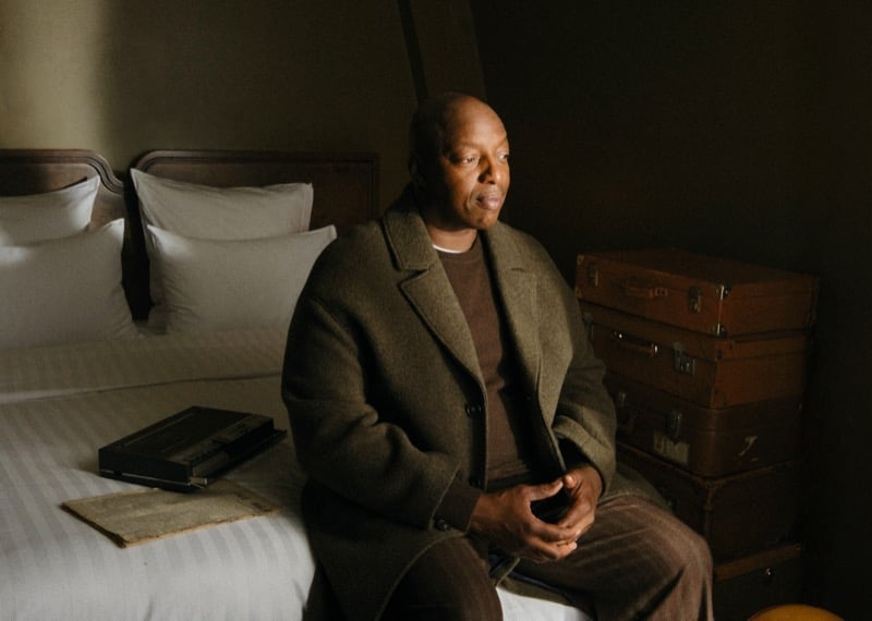 Oxmo Puccino seated on the edge of a hotel bed in a wool overcoat, hands clasped, beside a stack of vintage suitcases