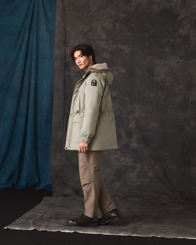 Model wears a gray-green Parajumpers coat and brown trousers against a dark studio backdrop.