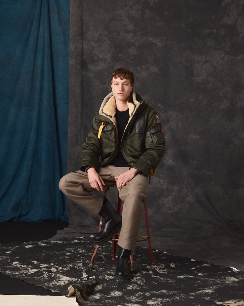 Seated model wears a Parajumpers Gobi jacket with layered knitwear and brown trousers.