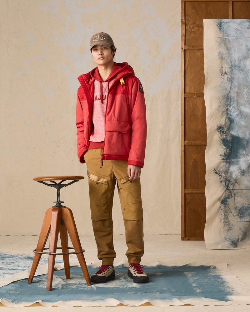 Model wears a red Parajumpers jacket and hoodie with ochre trousers and a cap.