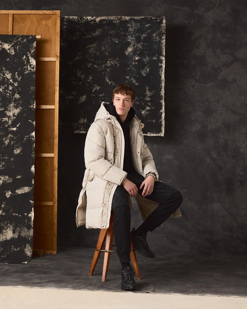 Model sits on a stool wearing a pale Parajumpers long coat and dark trousers.