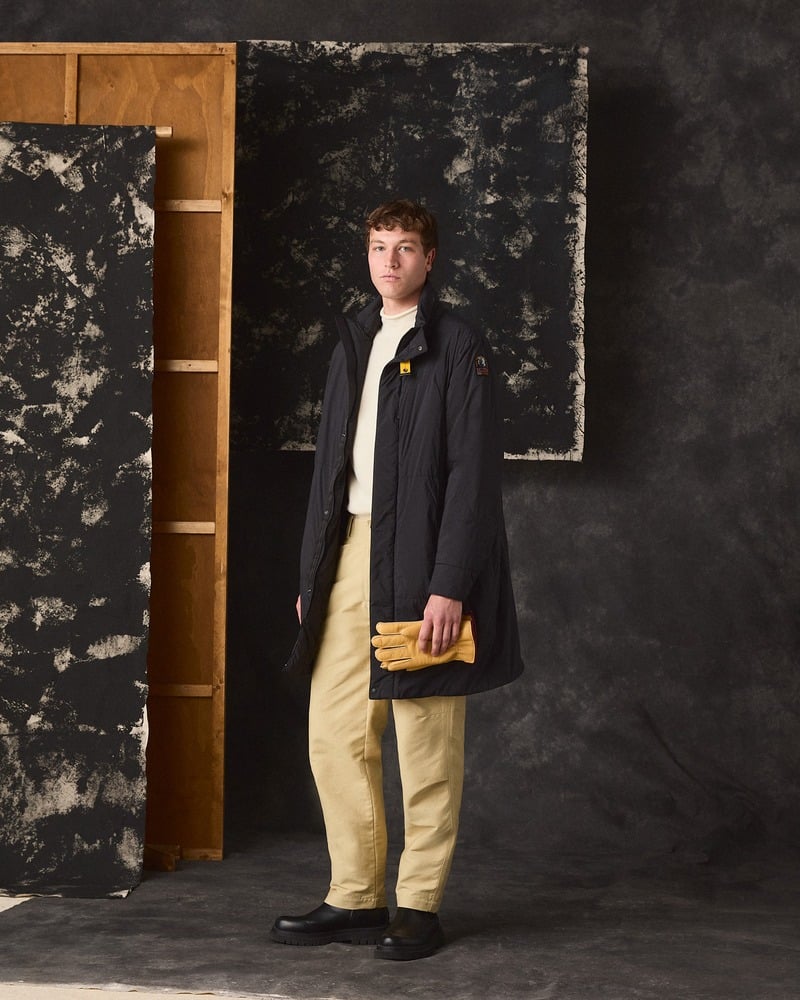 Model wears a dark Parajumpers long coat with tan trousers and carries yellow gloves.