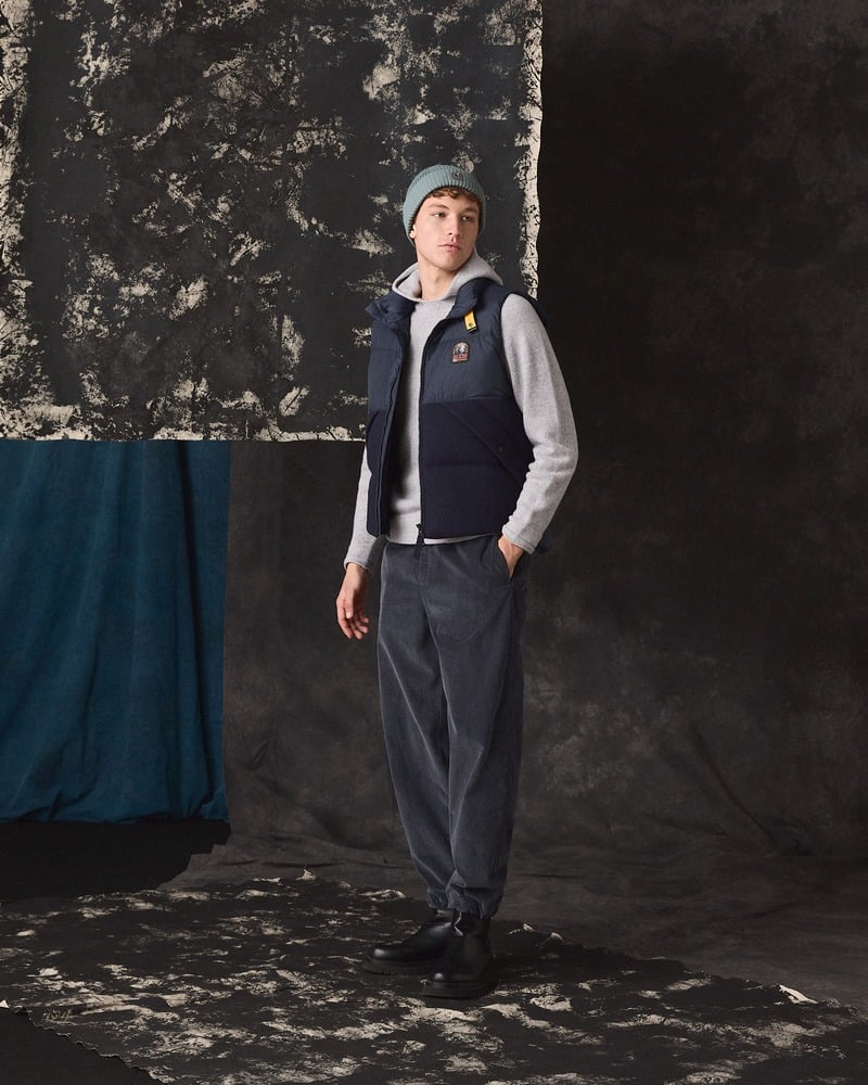 Model wears a navy Parajumpers padded vest over a gray sweater with wide trousers and a beanie.