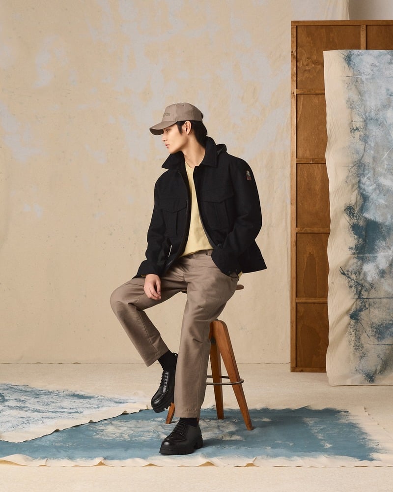 Seated model wears a dark Parajumpers jacket with brown trousers, boots, and a cap.