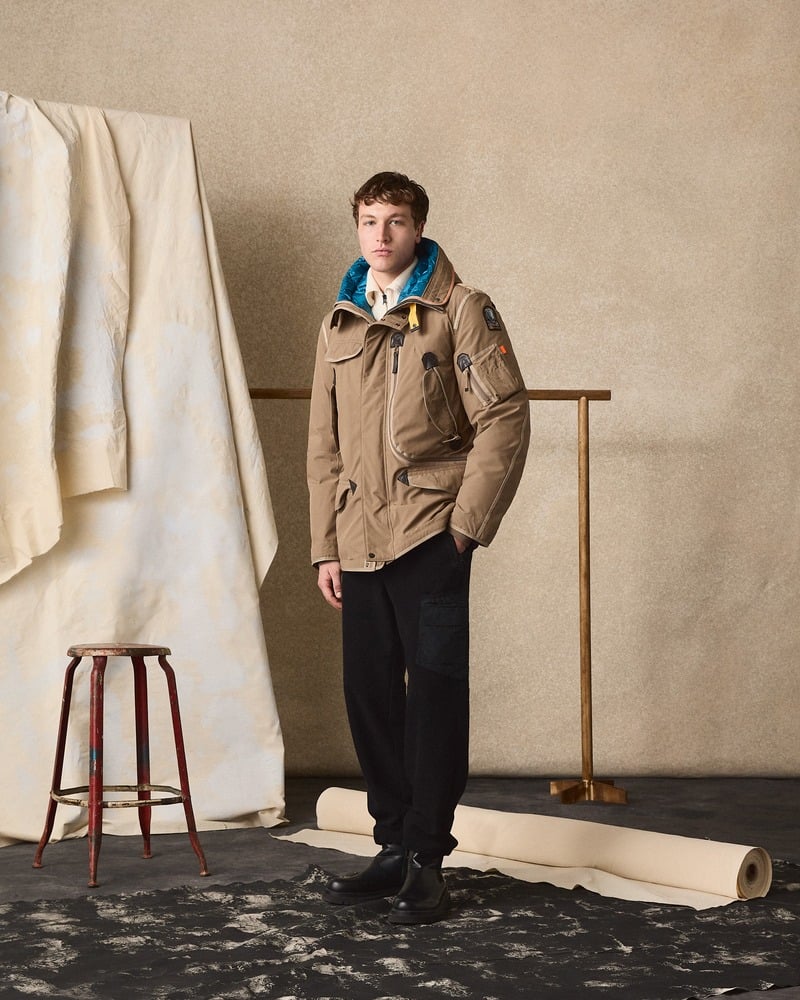 Model wears a brown Parajumpers hooded jacket with a blue layer and dark trousers.