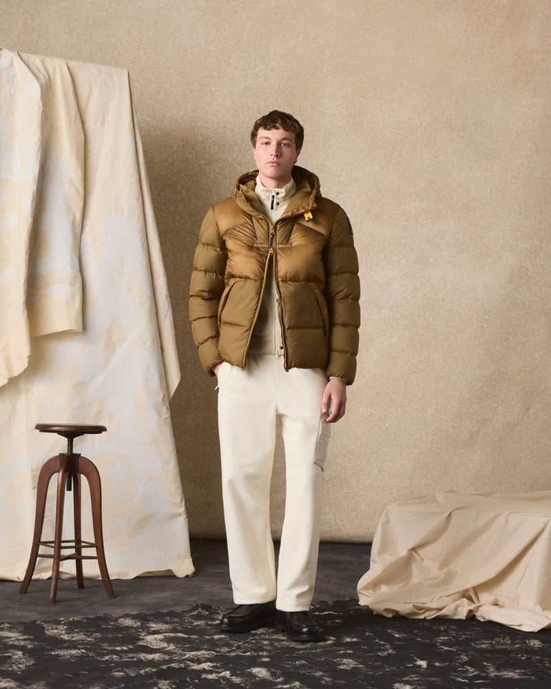 Model wears a brown Parajumpers padded jacket with white trousers in a studio.