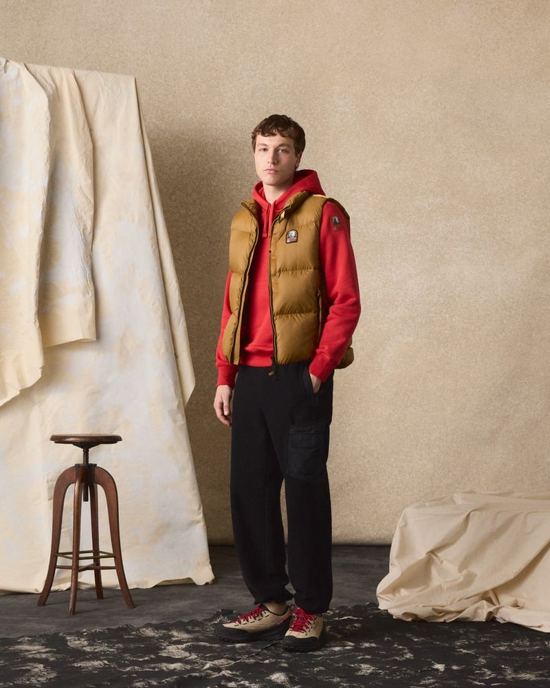 Model wears a brown Parajumpers padded vest over a red hoodie with dark trousers.
