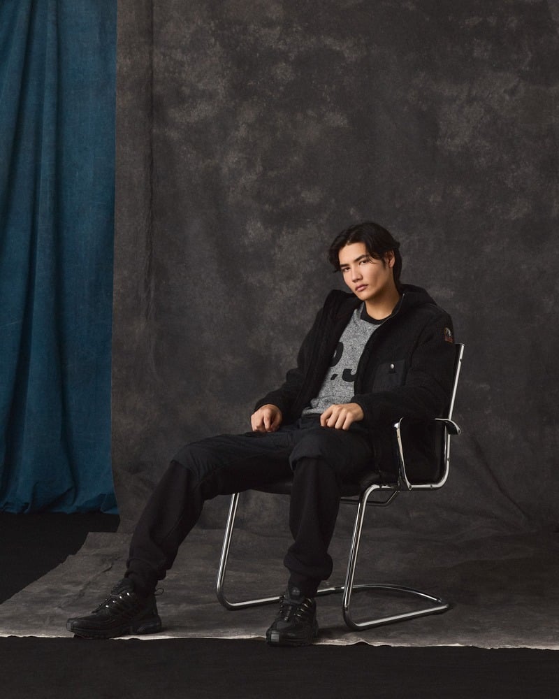 Model reclines in a metal chair wearing a dark Parajumpers outfit over a gray top.