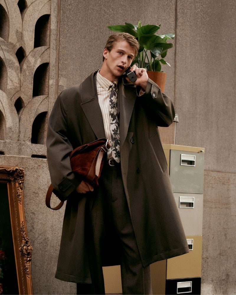 Vasko Luyckx holds a telephone receiver to his ear, wearing a long brown coat over a patterned shirt and tie.