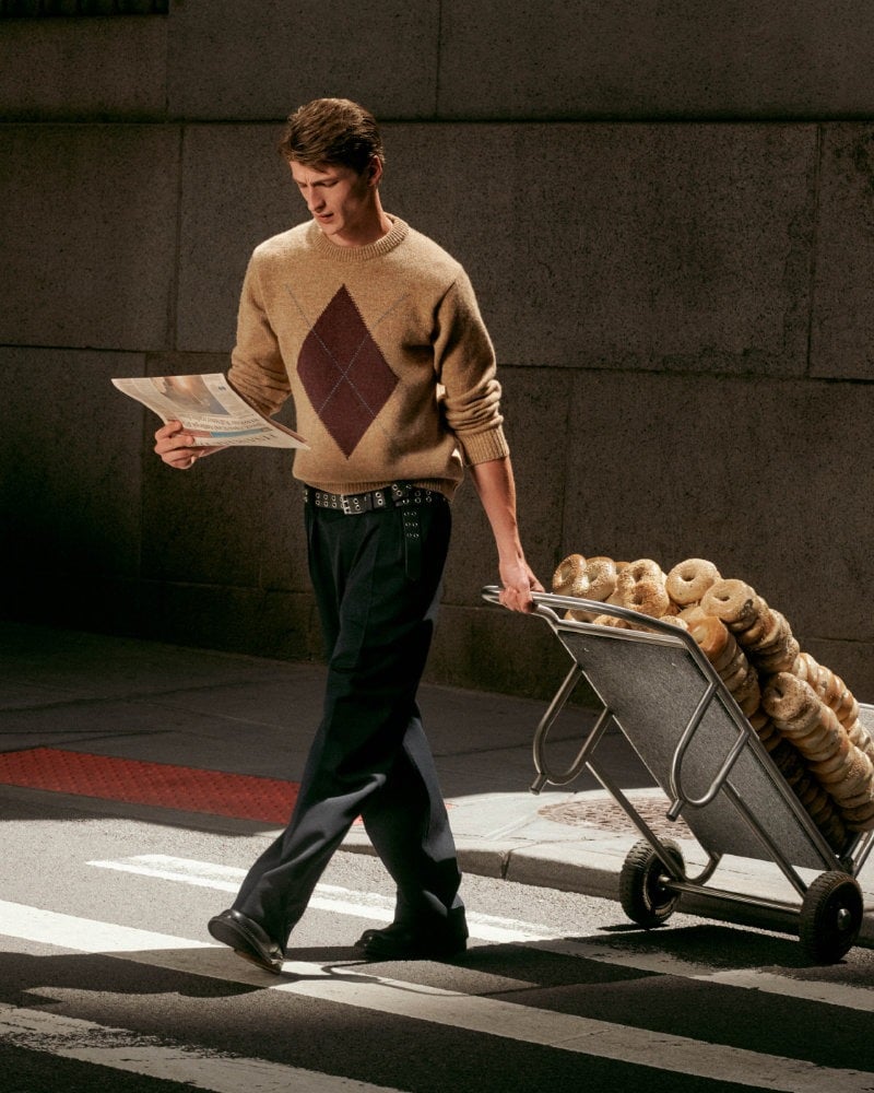 Vasko Luyckx pulls a trolley loaded with bagels, wearing a diamond-pattern sweater and dark trousers.