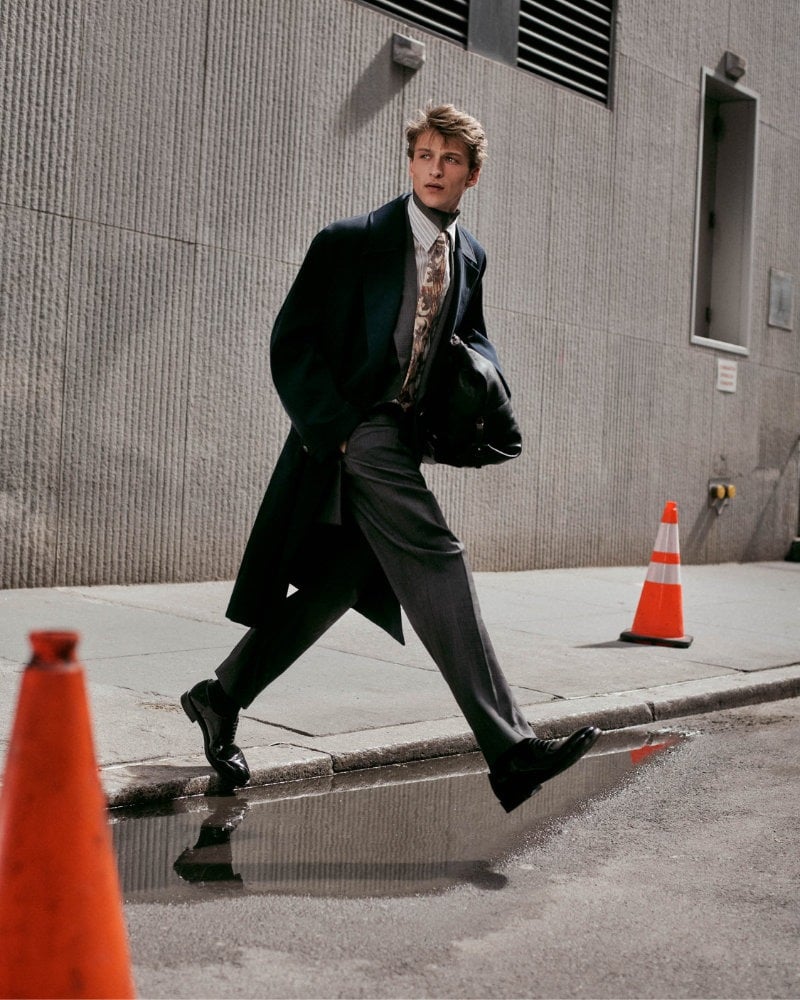 Vasko Luyckx leaps across a New York street in a long dark coat, shirt and tie, and full trousers.