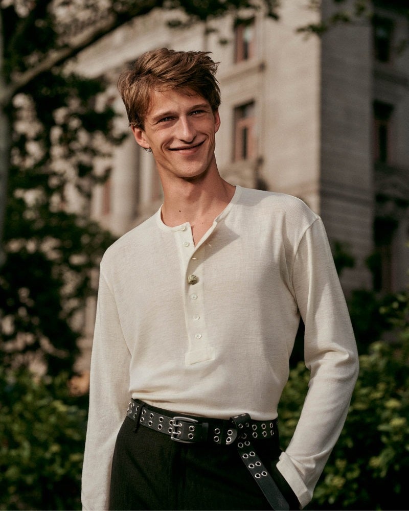Vasko Luyckx smiles in an ecru Henley tucked into dark trousers with an eyelet belt.