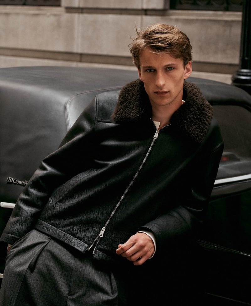 Vasko Luyckx leans against a car in a black leather jacket with a textured collar and pleated dark trousers.
