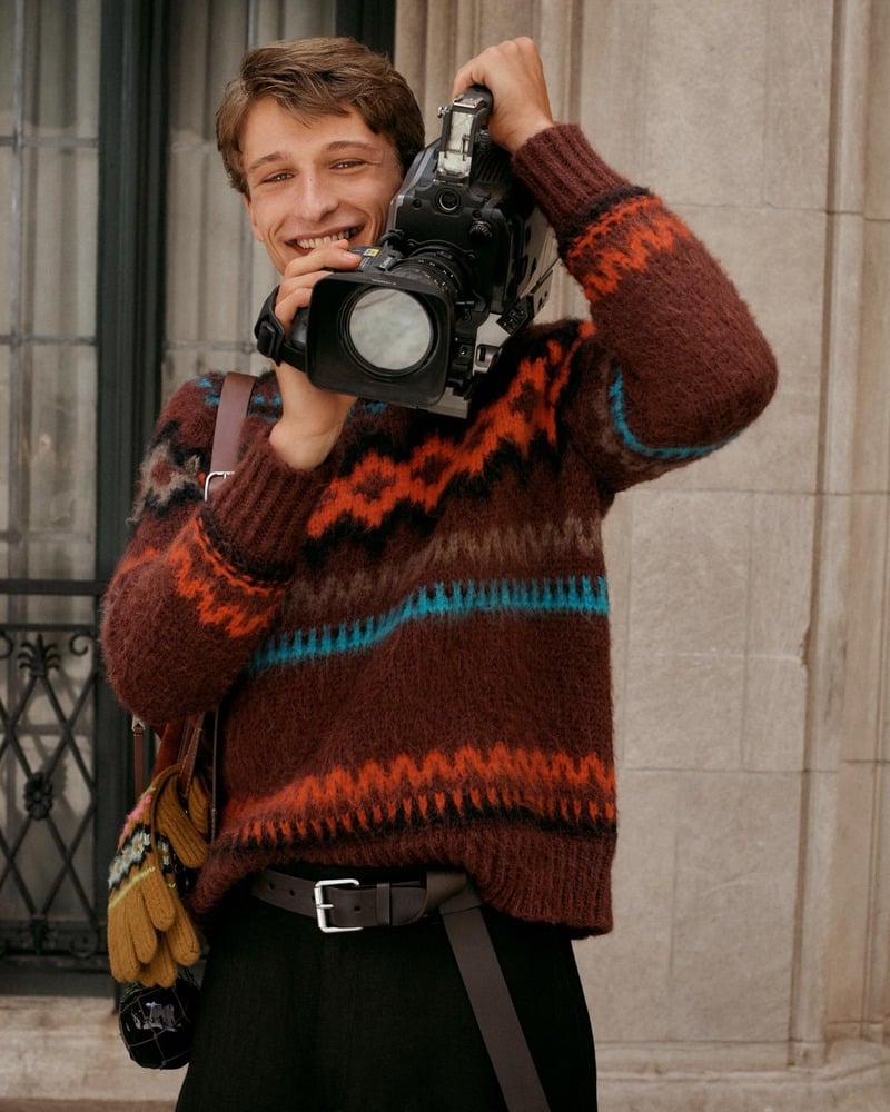 Vasko Luyckx holds a camera to his shoulder, wearing a colorful patterned sweater and dark trousers.