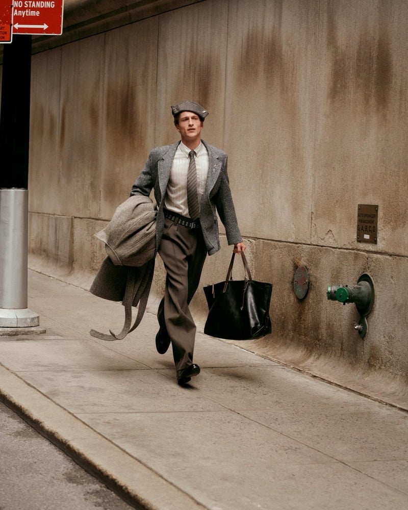 Vasko Luyckx walks along a sidewalk in a flat cap and tailored jacket, carrying a coat and dark bag.