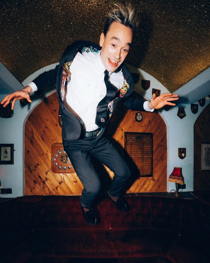 Ned Green of Legss leaps with arms outstretched, revealing a printed jacket lining and sunflower tie in the Paul Smith + Fender campaign.