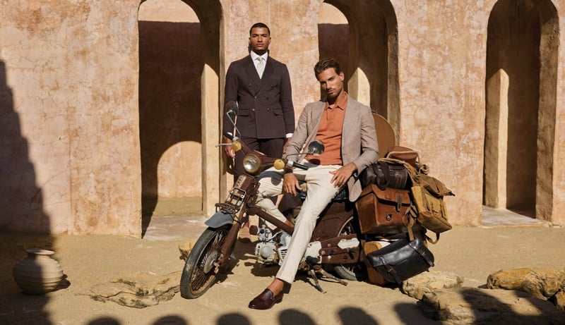 Paulo Lopes Lucio wears a dark double-breasted suit beside Gonçalo Campos, seated on a luggage-laden motorcycle in a taupe blazer and rust polo.