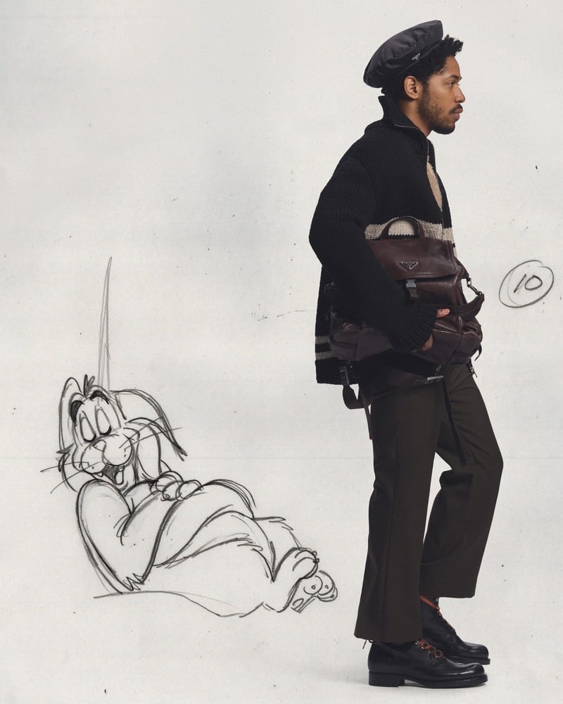 Kelvin Harrison Jr. wears a dark jacket, cropped trousers, and a cap with a brown bag behind the scenes of Prada's holiday 2026 campaign.