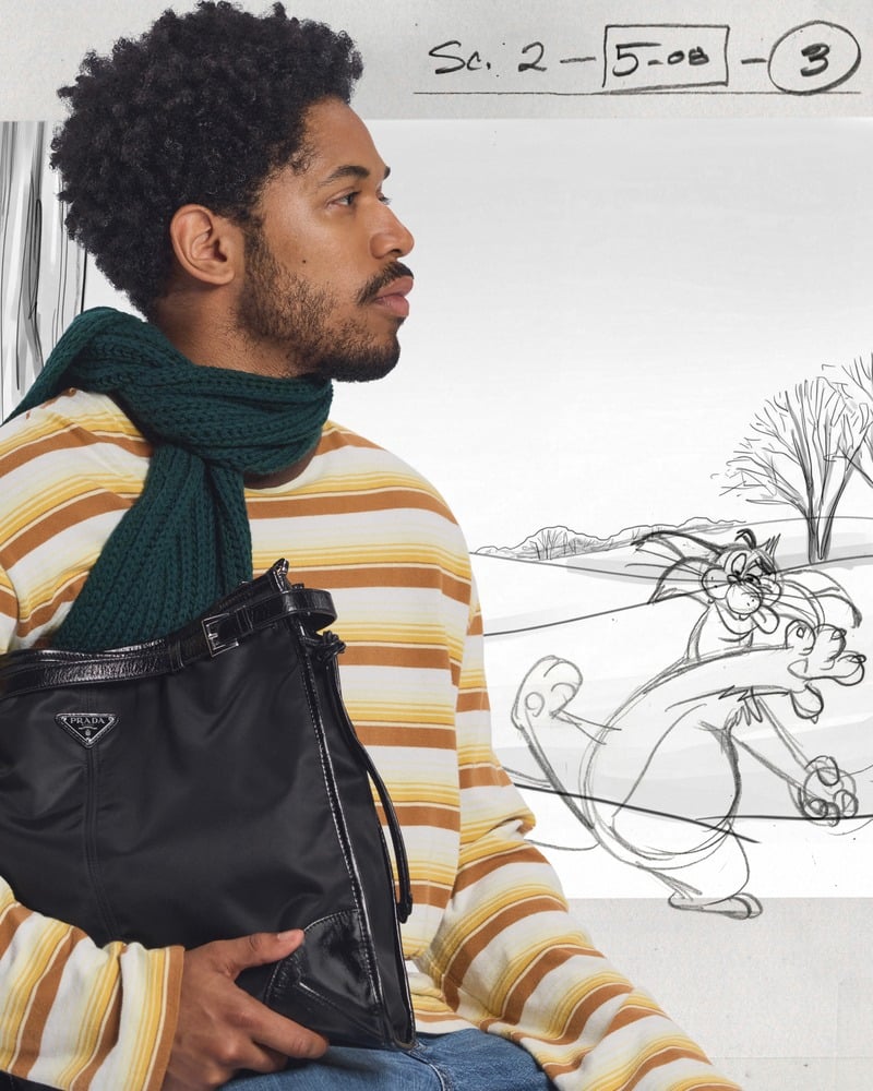 Kelvin Harrison Jr. wears yellow striped knitwear and a green scarf while holding a black bag behind the scenes of Prada's holiday 2026 campaign.