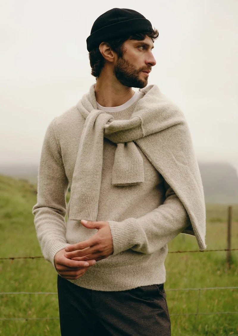 Matthew Bell wears an oatmeal sweater with a knit draped around his shoulders and a black beanie in a grassy field.