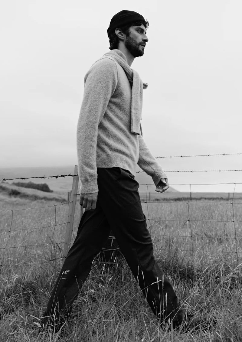 Matthew Bell walks through grass in a pale sweater and dark trousers in a black-and-white photograph.