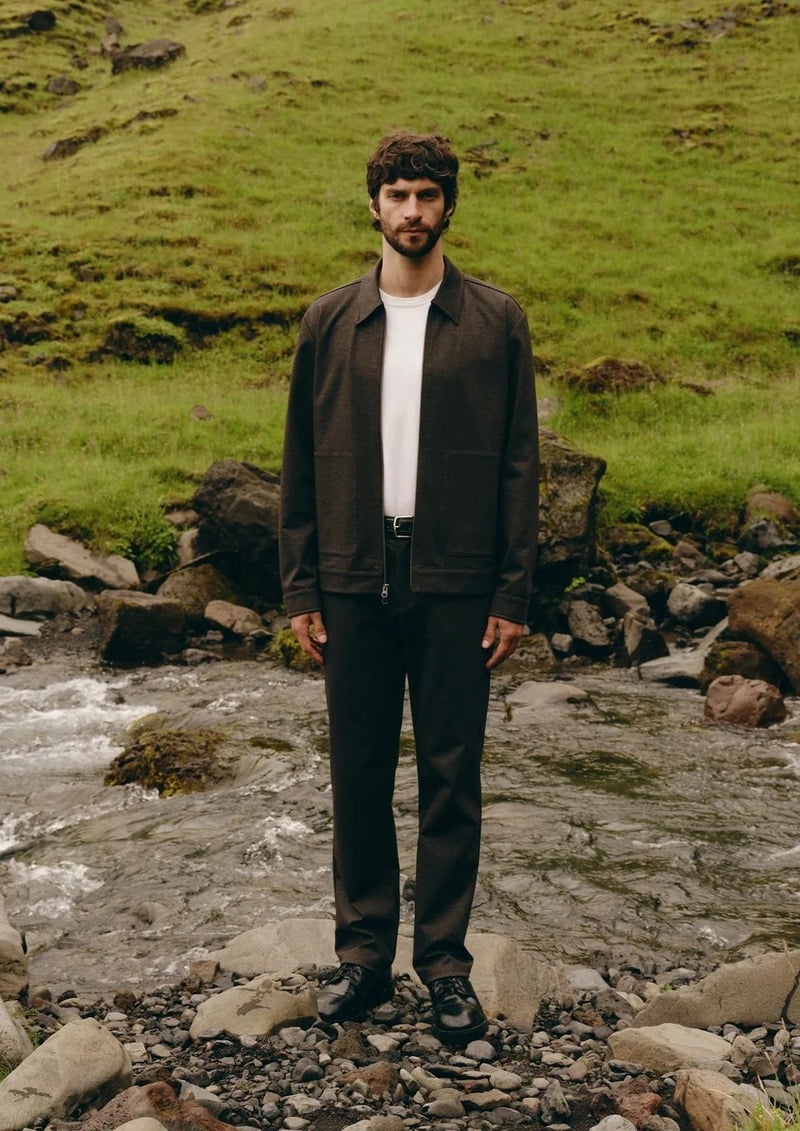 Matthew Bell stands beside a stream in a dark jacket, white T-shirt, and dark trousers.