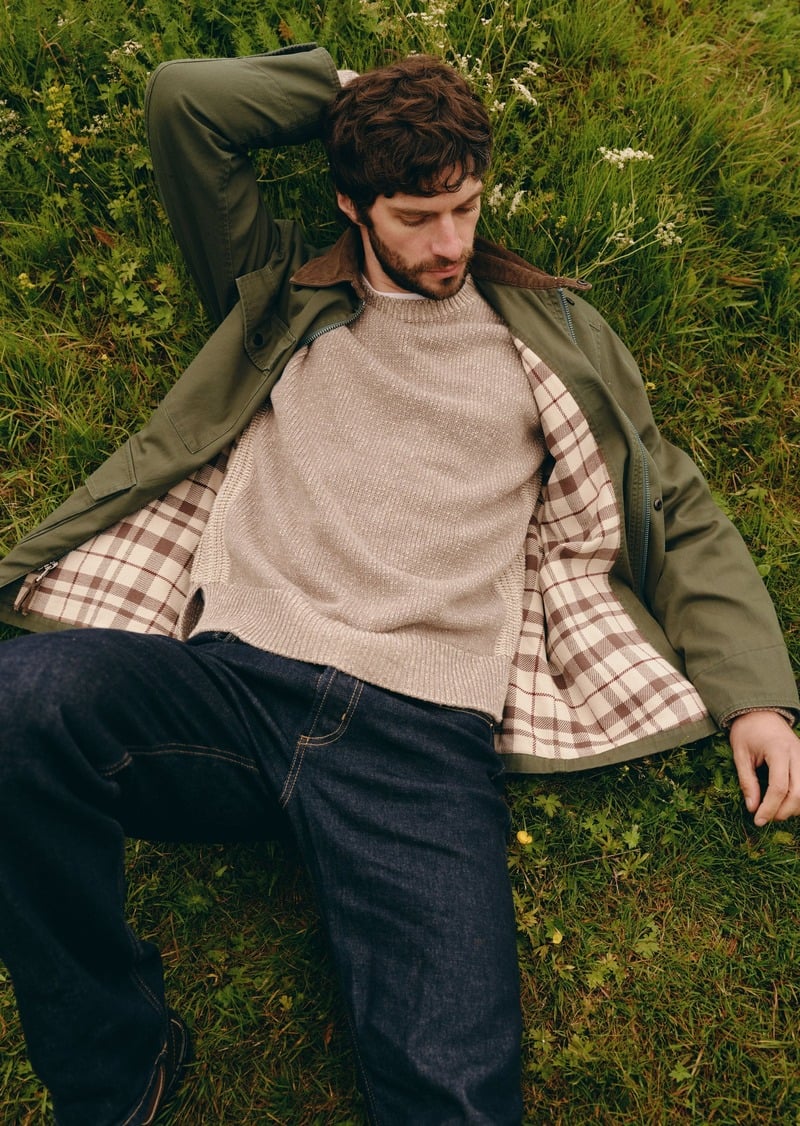 Matthew Bell reclines in the grass wearing an olive jacket with a checked lining and a pale crewneck.