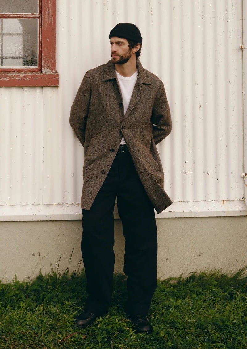 Matthew Bell wears a long brown coat over a white T-shirt and dark trousers beside a corrugated building.