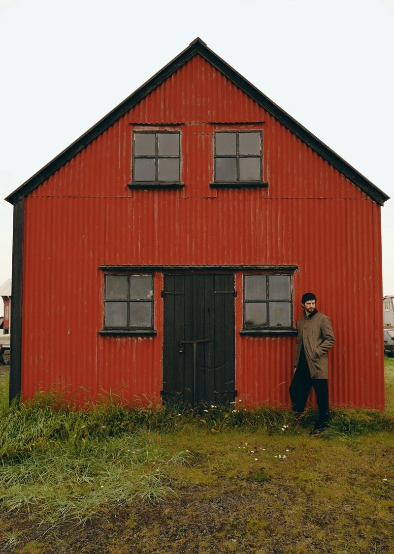 Matthew Bell stands beside a red house wearing a long brown coat and dark trousers.