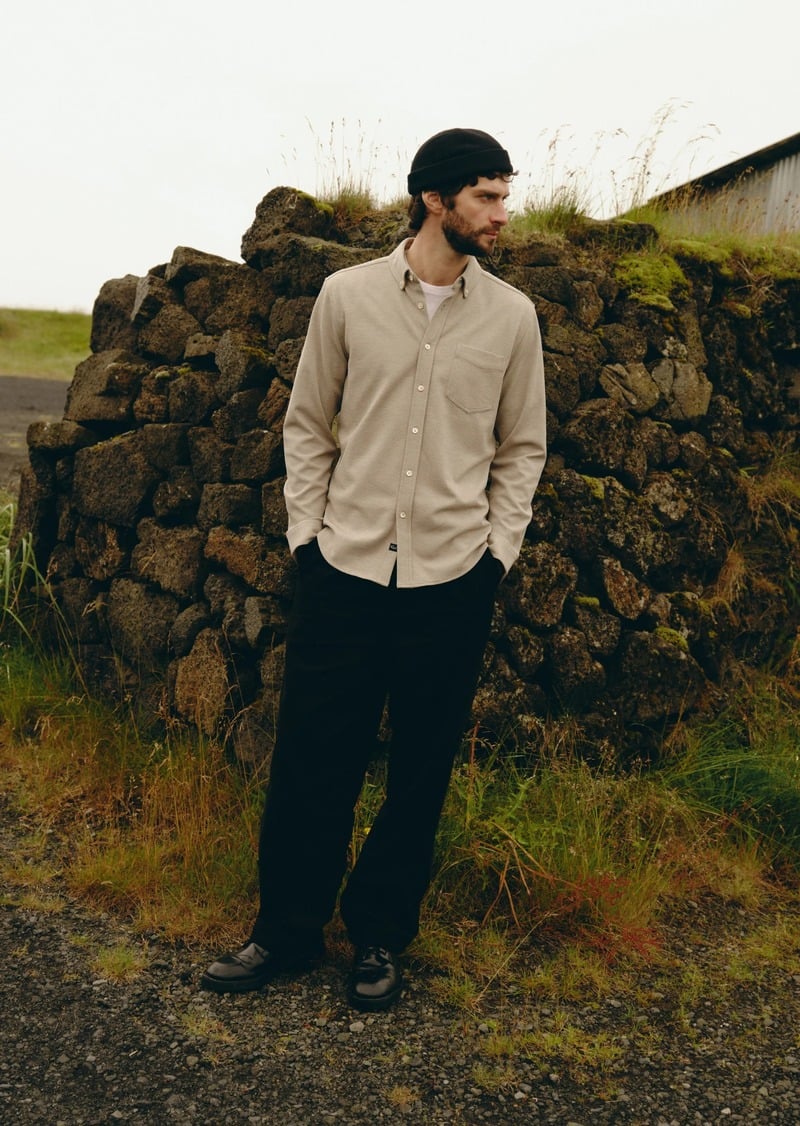 Matthew Bell wears a pale button-down shirt and dark trousers beside a stone wall.