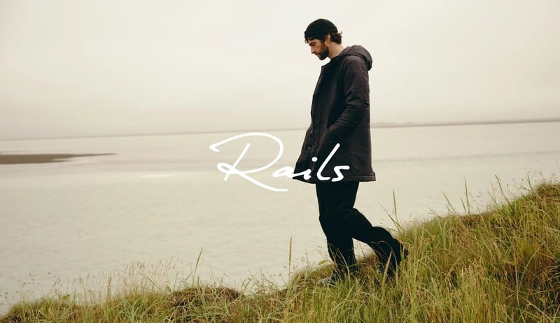 Matthew Bell walks beside the water in dark clothing beneath an overlaid Rails logo.