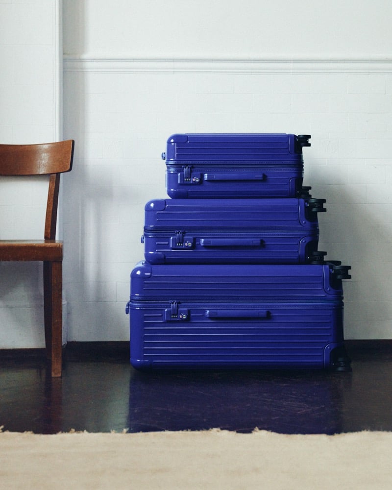 Three Ultramarine RIMOWA Essential suitcases stacked from large to small against a white brick wall beside a wooden chair