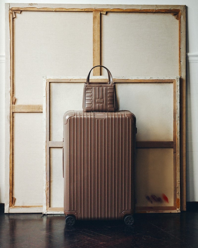 A brown RIMOWA Essential suitcase with a matching ribbed handbag on top, standing in front of two stretched canvases on a dark wood floor